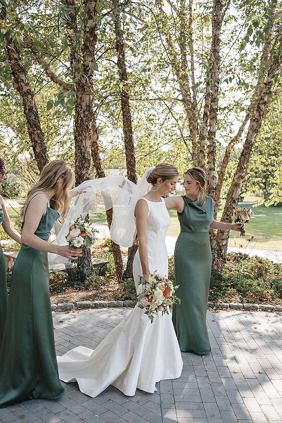 Bride with bridesmaids adjusting veil as she holds a bouquet, bridesmaids in green dresses on a birch-lined lawn path outdoors