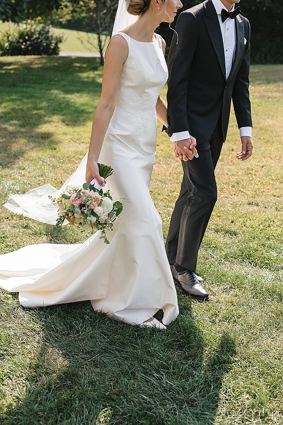 Couple portrait of bride and groom holding hands, walking on a sunlit lawn with trees, her veil and bouquet, his tuxedo boutonniere