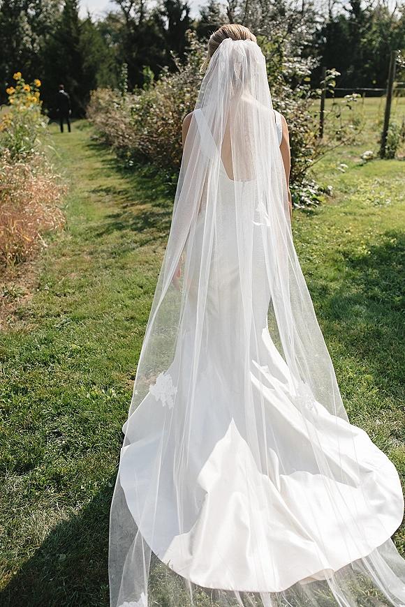 Bridal portrait of a back view bride in a cathedral veil, walking away in a sleeveless gown with train on a garden path outdoors
