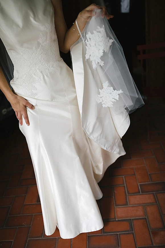 Wedding dress close-up showing a lace wedding dress bodice and tulle veil detail, with bracelet over terracotta tile floor by a dark doorway
