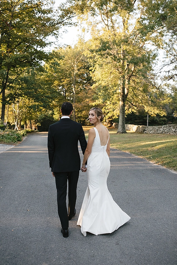 Couple portrait of bride and groom walking away hand in hand, bride in satin dress and groom in tuxedo on a sunlit tree-lined road