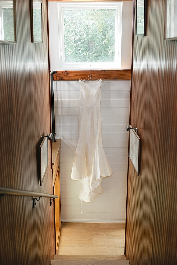 Wedding dress hanging on a hanger in soft window light, framed by wooden walls and a staircase with trees visible outside