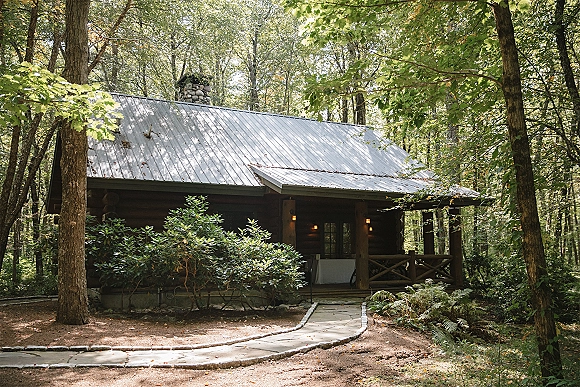 Forest cabin venue with a log cabin, stone chimney, and porch lantern lights along a stone walkway surrounded by woods