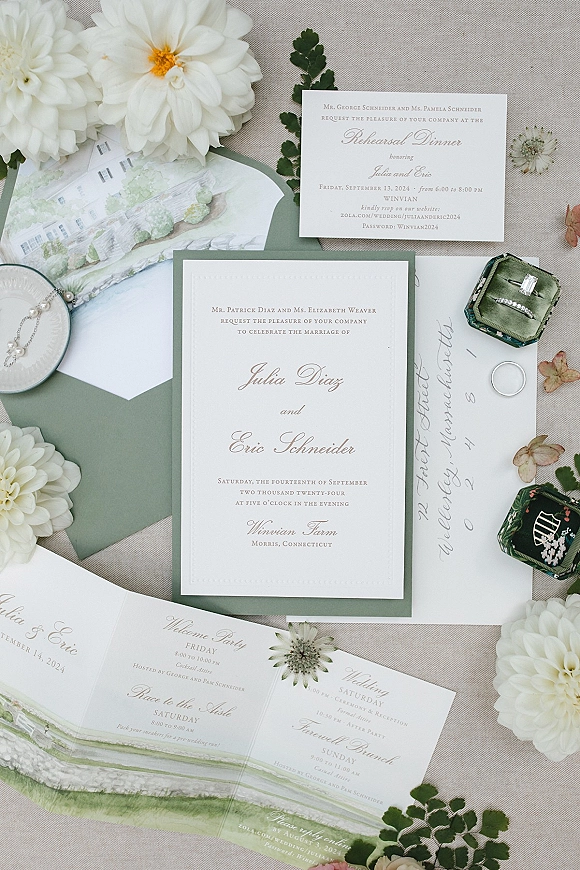 Wedding invitation suite with green and white invitations, sage envelopes, wax seal, rings and pearl bracelet on a linen backdrop with dahlias