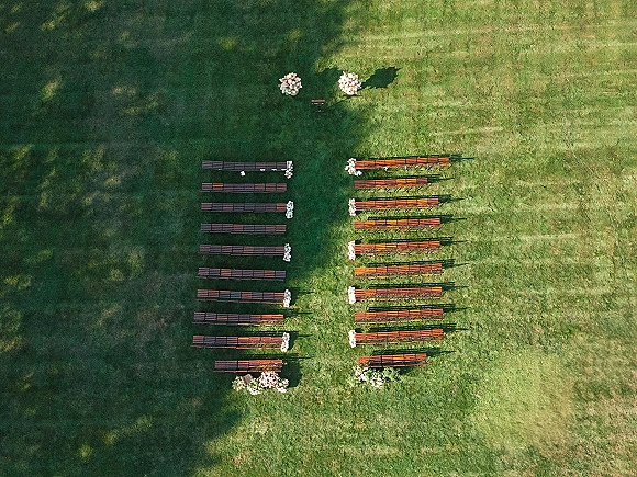 Ceremony setup with outdoor ceremony seating arranged symmetrically around an aisle runner, wood benches and floral arrangements on a grassy lawn with tree shadows