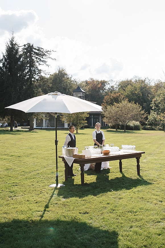 Outdoor bar setup wedding cocktail station with wooden table, white umbrella, champagne bucket, glassware and dispensers on a lawn with trees