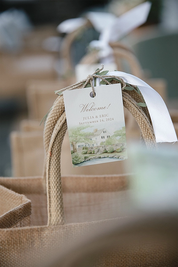 Wedding welcome tag tied with twine on a burlap tote welcome gift bag, finished with white ribbon and a small greenery sprig indoors