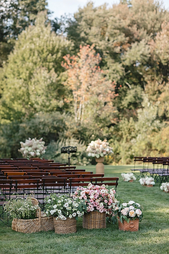 Outdoor ceremony setup with garden wedding ceremony aisle flowers in woven baskets and urns lining wooden benches on a green lawn