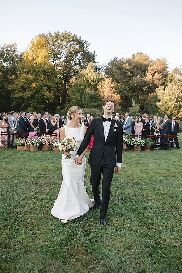 Recessional moment as bride and groom walking hand in hand, laughing past guests on a grassy aisle, bouquet in hand under trees