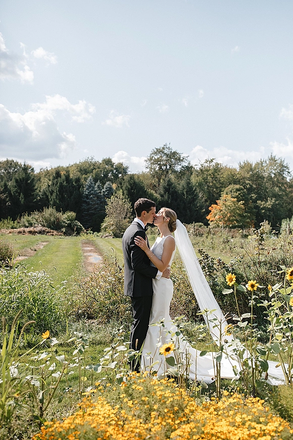 Wedding kiss portrait of bride and groom kissing, her veil flowing over a wildflower field path under blue sky with clouds