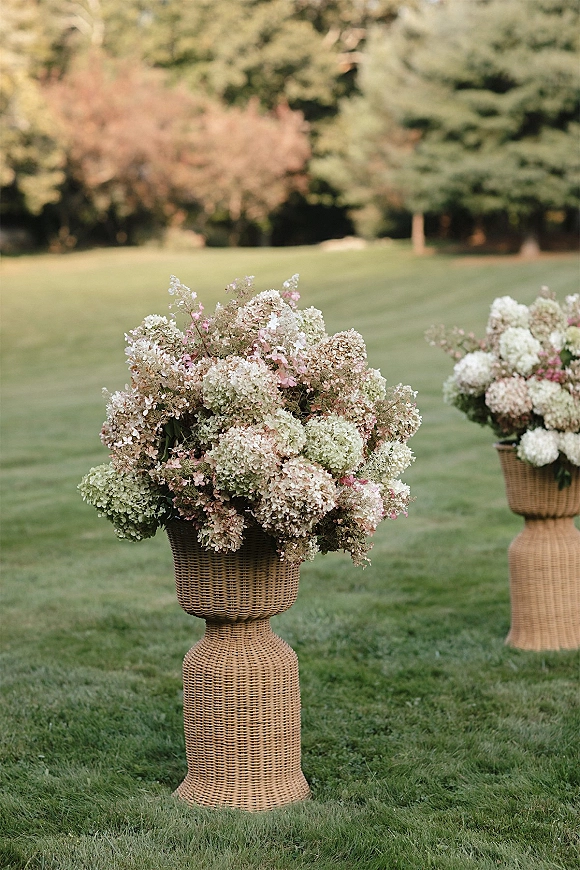 Ceremony floral arrangements of white and blush hydrangea aisle flowers on wicker pedestals in a garden lawn with trees backdrop