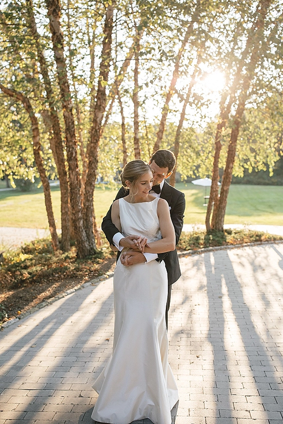 Couple portrait of bride and groom embrace on a paved path, groom hugging bride in a wedding dress as sun flares through trees