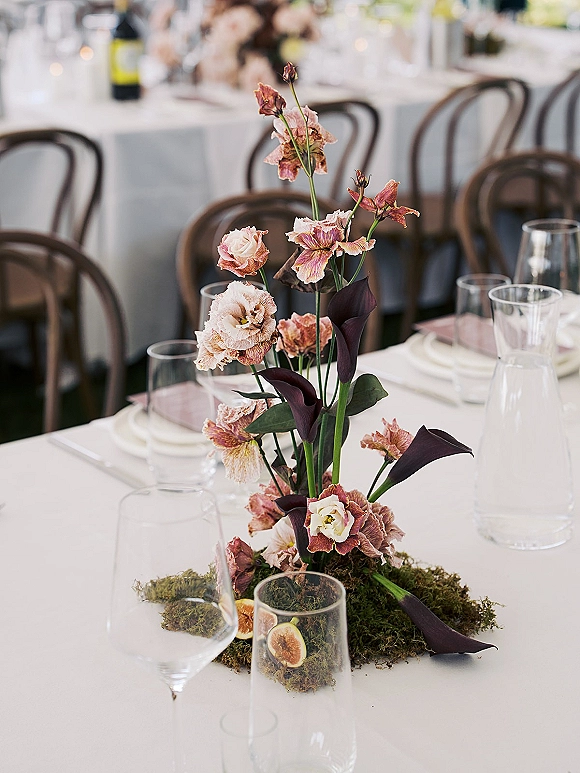 Wedding centerpiece with a calla lily centerpiece of blush flowers on a moss runner, with figs, glassware, and bentwood chairs in soft focus