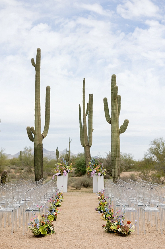 Ceremony aisle design with colorful wildflower aisle florals lining a sand path, flanked by clear acrylic chairs and saguaro cacti under blue sky