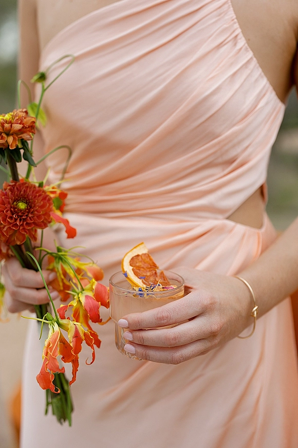 Bridesmaid dress in peach ruched satin with cutout waist, holding orange bouquet and citrus-garnished cocktail against blurred greenery backdrop