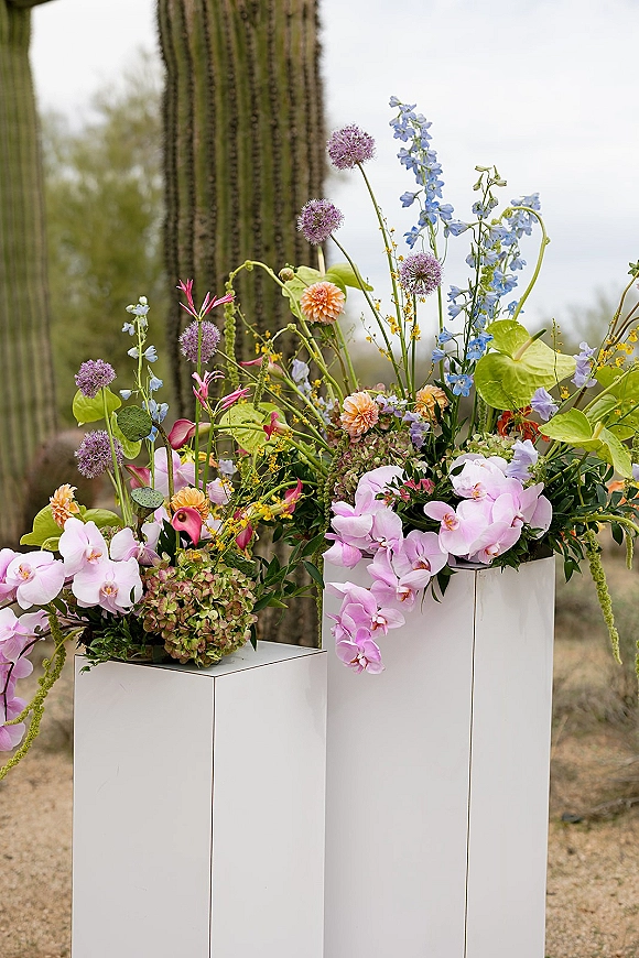 Wedding floral arrangement on white plinths with orchids, wildflowers, hydrangea and calla lilies, set against a desert cactus landscape