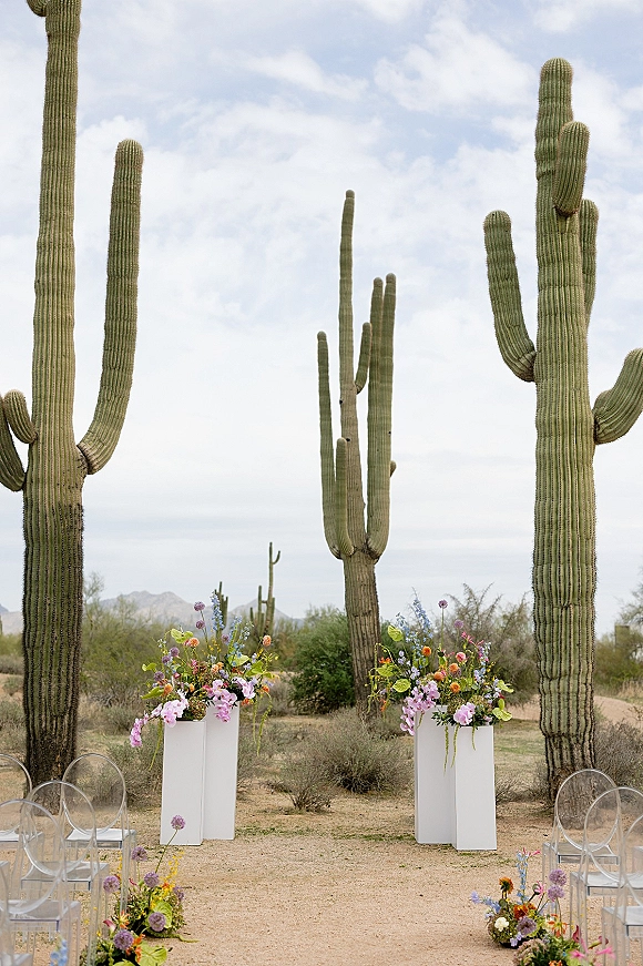 Ceremony altar decor with two white pedestal plinths and floral arrangements, clear acrylic chairs, and aisle flowers in a cactus desert landscape