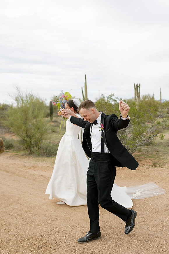 Wedding recessional as bride and groom walk away on a desert path, groom fist pumping while her veil trails behind, holding bouquet
