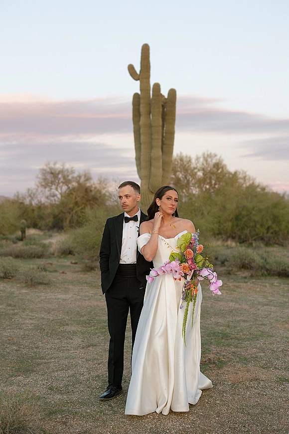 Couple portrait in a desert wedding portrait, bride in a strapless dress holding a colorful bouquet beside groom in tuxedo at sunset saguaro cactus backdrop