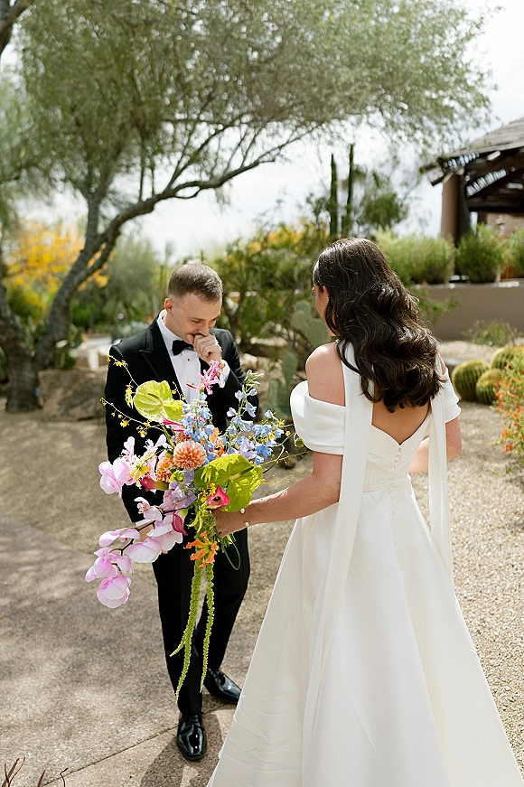 First look moment as groom in black tuxedo turns toward bride in off-shoulder gown holding a colorful bouquet on a desert garden path