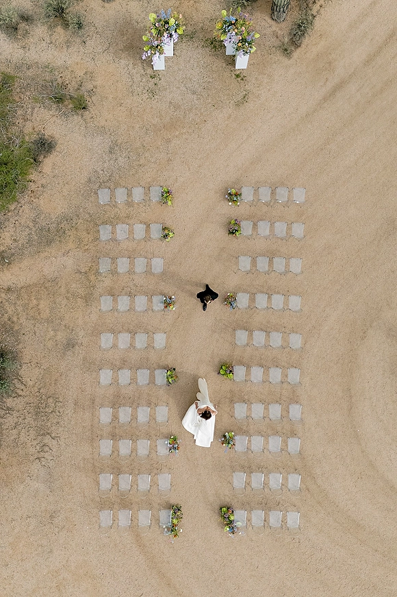 Ceremony setup with an outdoor ceremony aisle lined by aisle floral arrangements and rows of chairs on dirt ground among trees and shrubs