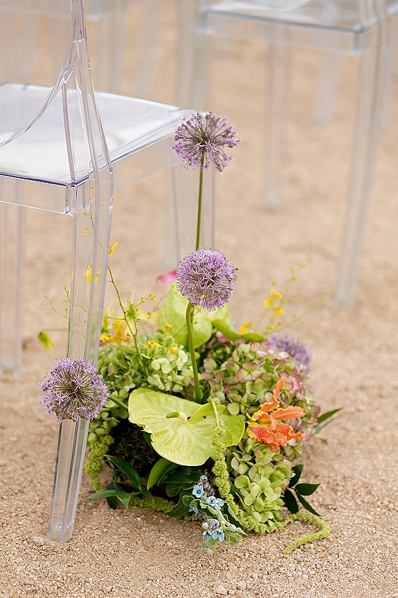 Aisle flower arrangement with wedding aisle flowers in purple allium, green anthurium, and hydrangea on gravel beside clear acrylic chairs
