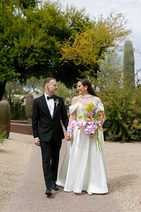 Couple portrait of bride and groom walking hand in hand, her off-the-shoulder dress with bouquet on a desert garden path with cactus