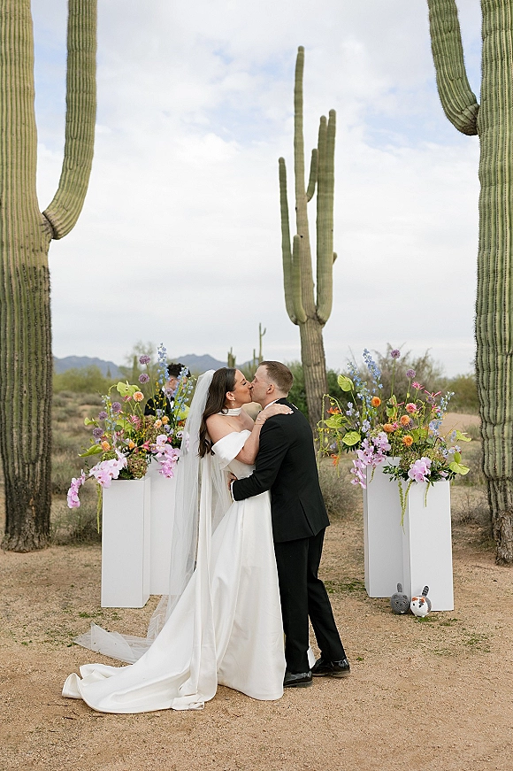 Wedding kiss portrait of bride and groom kissing under a long veil beside colorful florals on white pedestals in a saguaro desert landscape