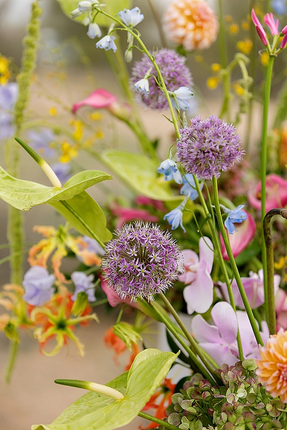 Wedding florals in a wildflower wedding flowers arrangement with purple allium, bluebells, dahlias, orchids, and lush greenery against a soft garden blur