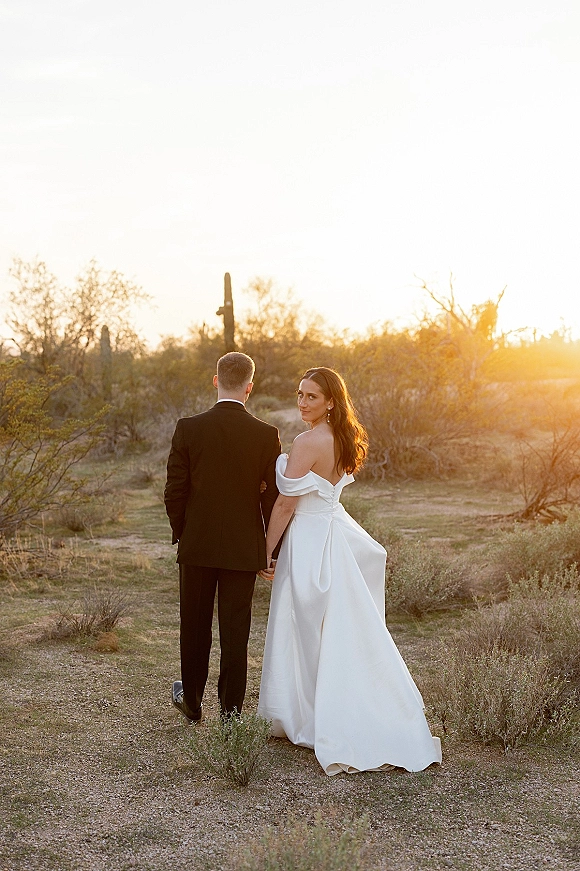 Couple portrait of bride and groom walking away holding hands on a desert path at sunset, bride glancing over shoulder in off-shoulder gown