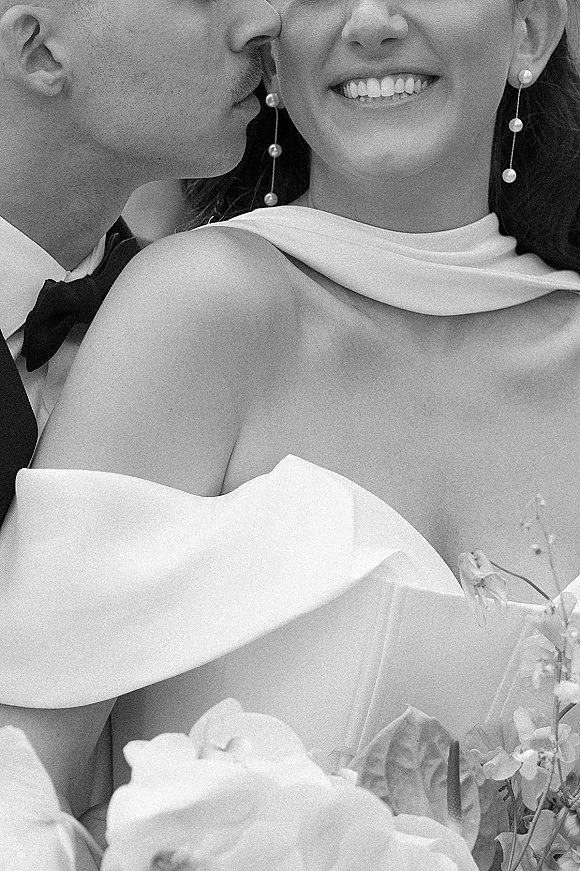 Wedding couple portrait in black and white with groom kissing the bride’s cheek as she smiles, wearing pearl drop earrings against a neutral backdrop