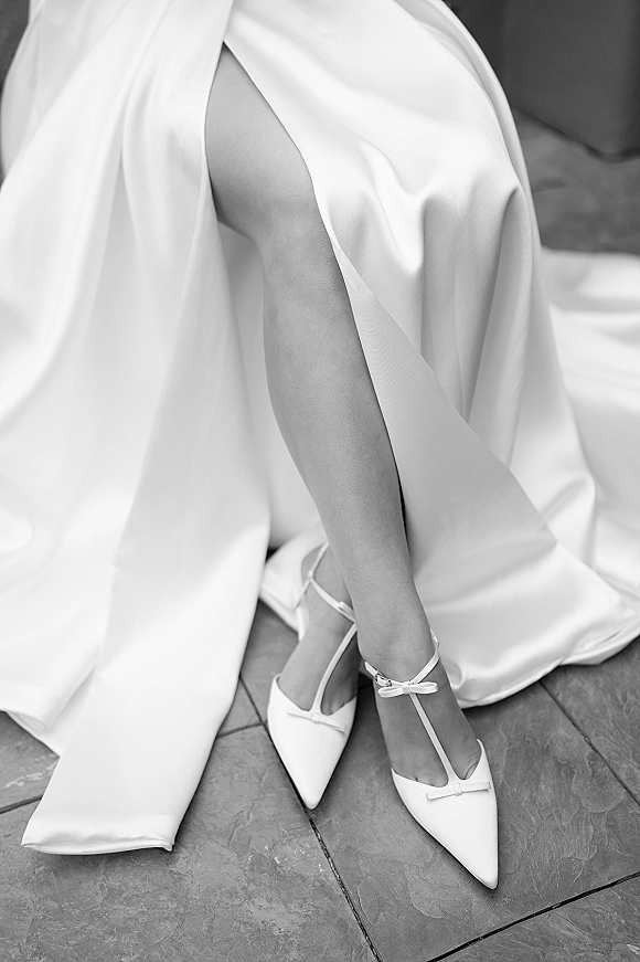Bridal shoes, white wedding heels with pointed toes and ankle-strap bows on a stone tile floor beside a sofa and satin dress hem