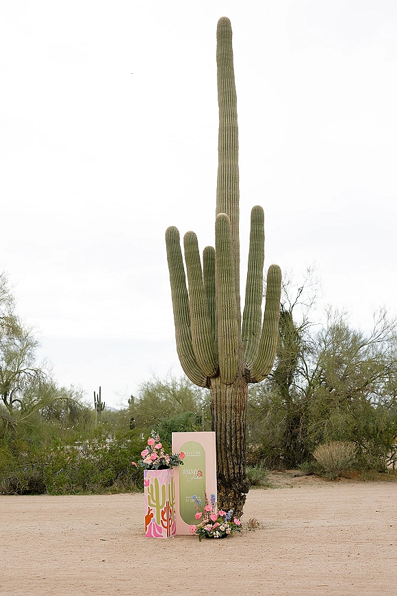 Wedding welcome sign on a pedestal plinth with a floral arrangement and cactus illustration, set against a saguaro desert landscape