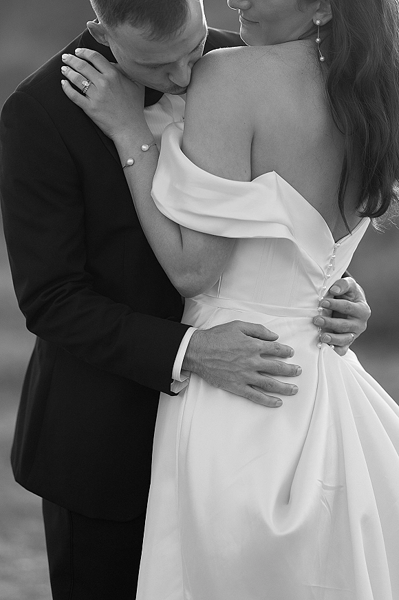 Wedding couple portrait of groom kissing bride’s shoulder as she shows her engagement ring, wearing pearl drop earrings by blurred shoreline