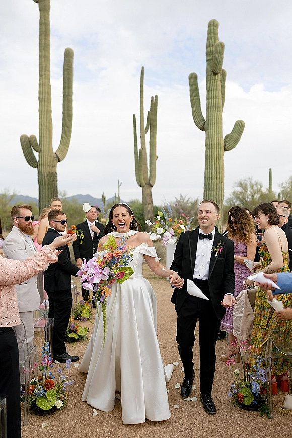 Wedding recessional as bride and groom walk hand in hand down a dirt aisle, petals flying, with saguaro cacti and mountains behind