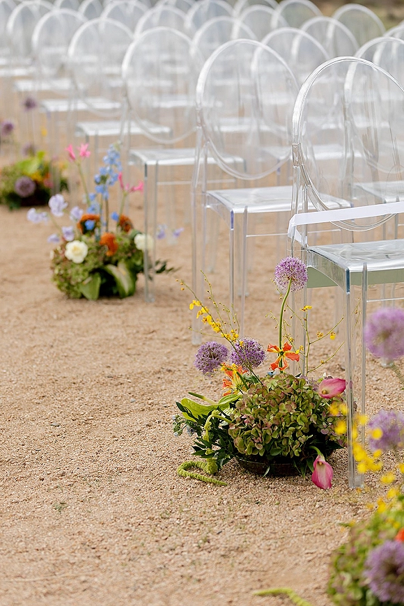 Ceremony aisle decor with outdoor ceremony aisle flowers lining a gravel path beside clear acrylic chairs tied with ribbon and florals