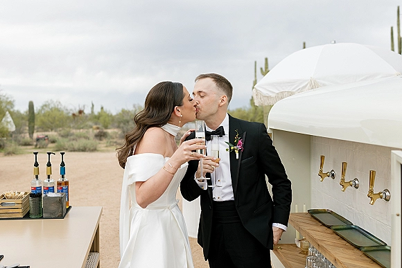 Wedding kiss as the bride and groom toast with champagne flutes beside a bar cart in a desert cactus landscape under cloudy skies