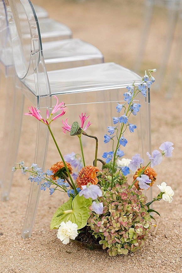 Ceremony aisle decor with acrylic ceremony chairs beside low wildflower clusters of blue delphinium, pink and orange blooms on a gravel aisle