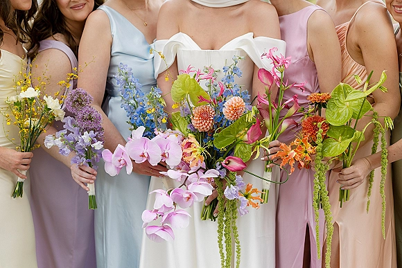 Bridesmaid bouquets in colorful wedding bouquets with cascading orchids, dahlias, and anthurium held by women in pastel dresses on a neutral backdrop
