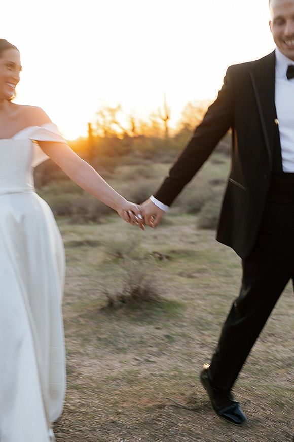 Couple portrait of bride and groom holding hands in a desert landscape at sunset, her off-the-shoulder gown and his tuxedo backlit