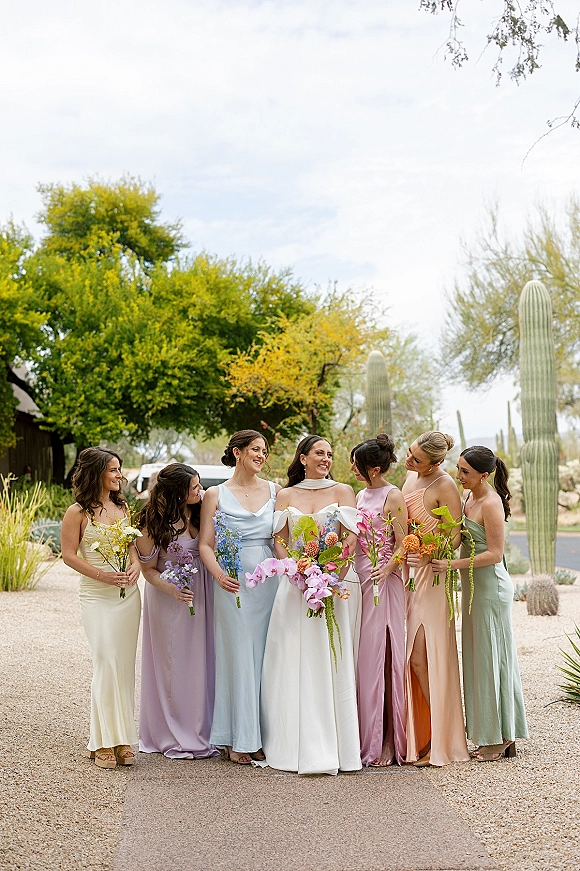 Bridesmaids portrait with bride in off-the-shoulder gown, pastel dresses, and orchid wildflower bouquets on a desert path with cactus backdrop