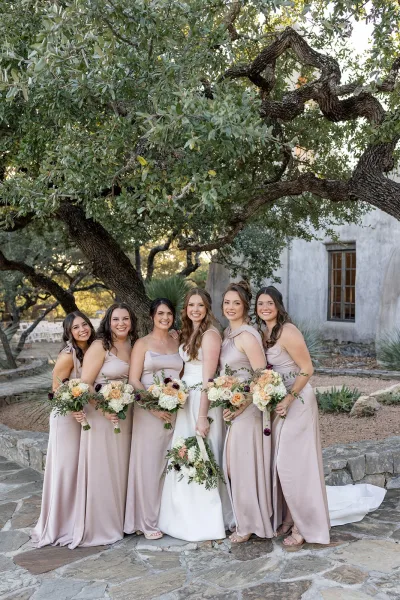 Bridesmaid group photo with bride with bridesmaids in neutral satin dresses holding rose and greenery bouquets on a stone patio by an oak tree