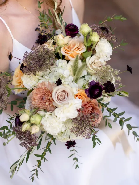 Bridal bouquet, garden style bridal bouquet with roses, ranunculus, dahlia and berry sprigs held at the bride’s torso against a neutral wall
