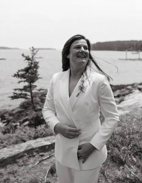 Bridal portrait of a bride in a white suit on a rocky waterfront dock, with trees along the shore beneath a wide sky.