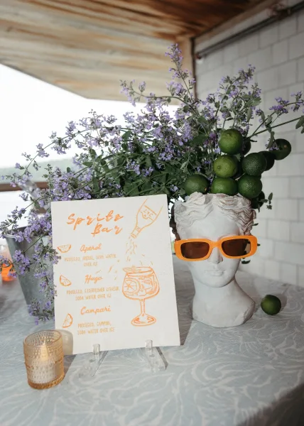 Wedding bar sign anchors a spritz bar setup, an illustrated cocktail sign styled with fresh limes on a table against a white tile wall.
