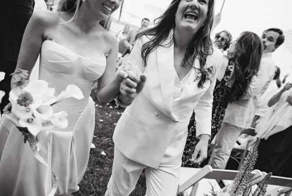 Wedding recessional with a couple holding hands as two brides walk, one in a white bridal suit, past cheering guests down the aisle.