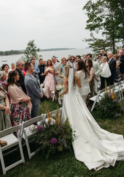 Wedding processional with bride walking down the aisle holding a bridal bouquet by a lakeside lawn under a cloudy sky