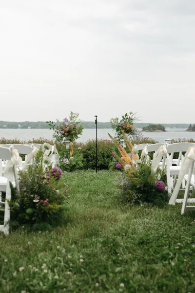 Outdoor ceremony setup with waterfront ceremony decor and white chairs on a grass lawn beside still water and distant trees under clouds.