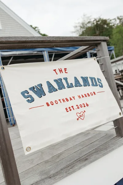 Wedding sign styled as a personalized wedding banner with a heart motif, hanging on a wood deck railing by a building and trees under overcast sky.