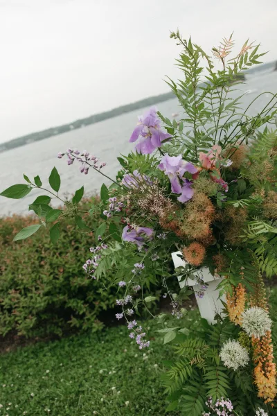 Ceremony flowers styled as lush aisle flower arrangements on a white pedestal, set beside a lake with shrubs and a grassy lawn under clouds.